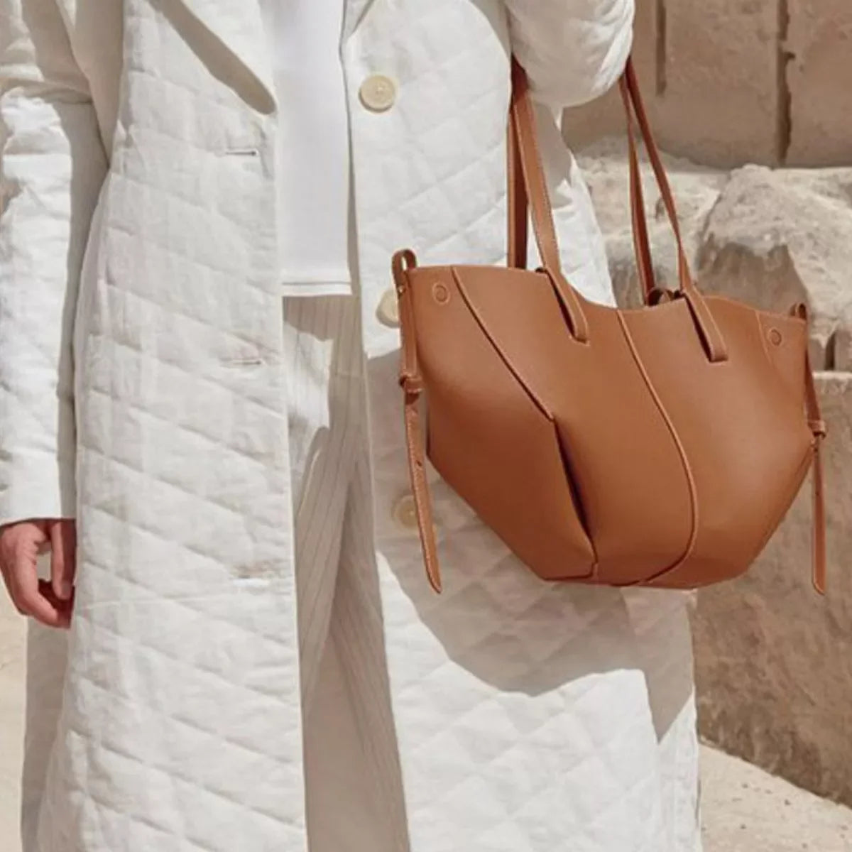 Person wearing white quilted coat holding a brown leather tote bag outdoors against stone wall