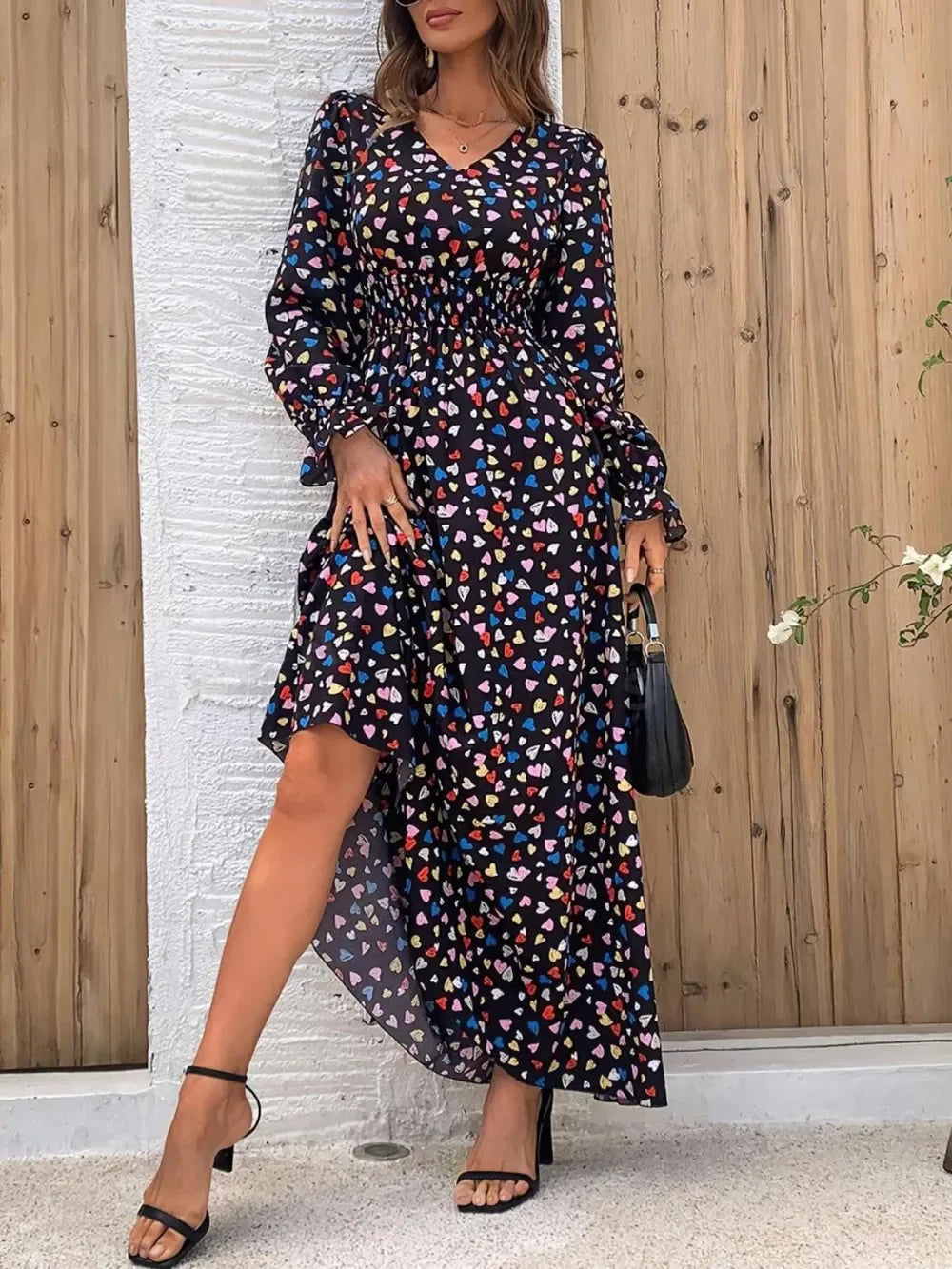 Woman wearing black long-sleeve maxi dress with colorful heart prints, standing by wooden and white walls