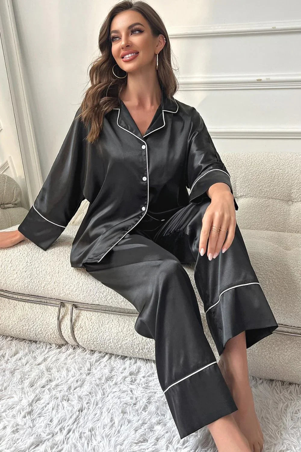 Woman sitting on white sofa wearing black satin pajamas with white piping, smiling indoors