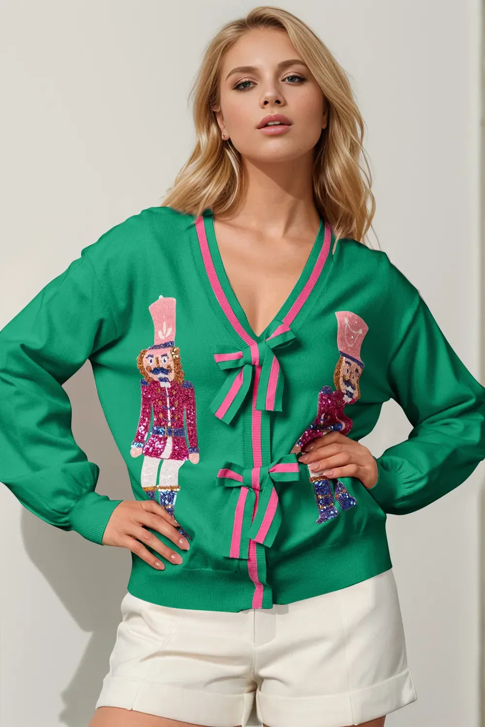 Blonde woman wearing green cardigan with pink ribbon ties and nutcracker soldier sequin designs, paired with white shorts