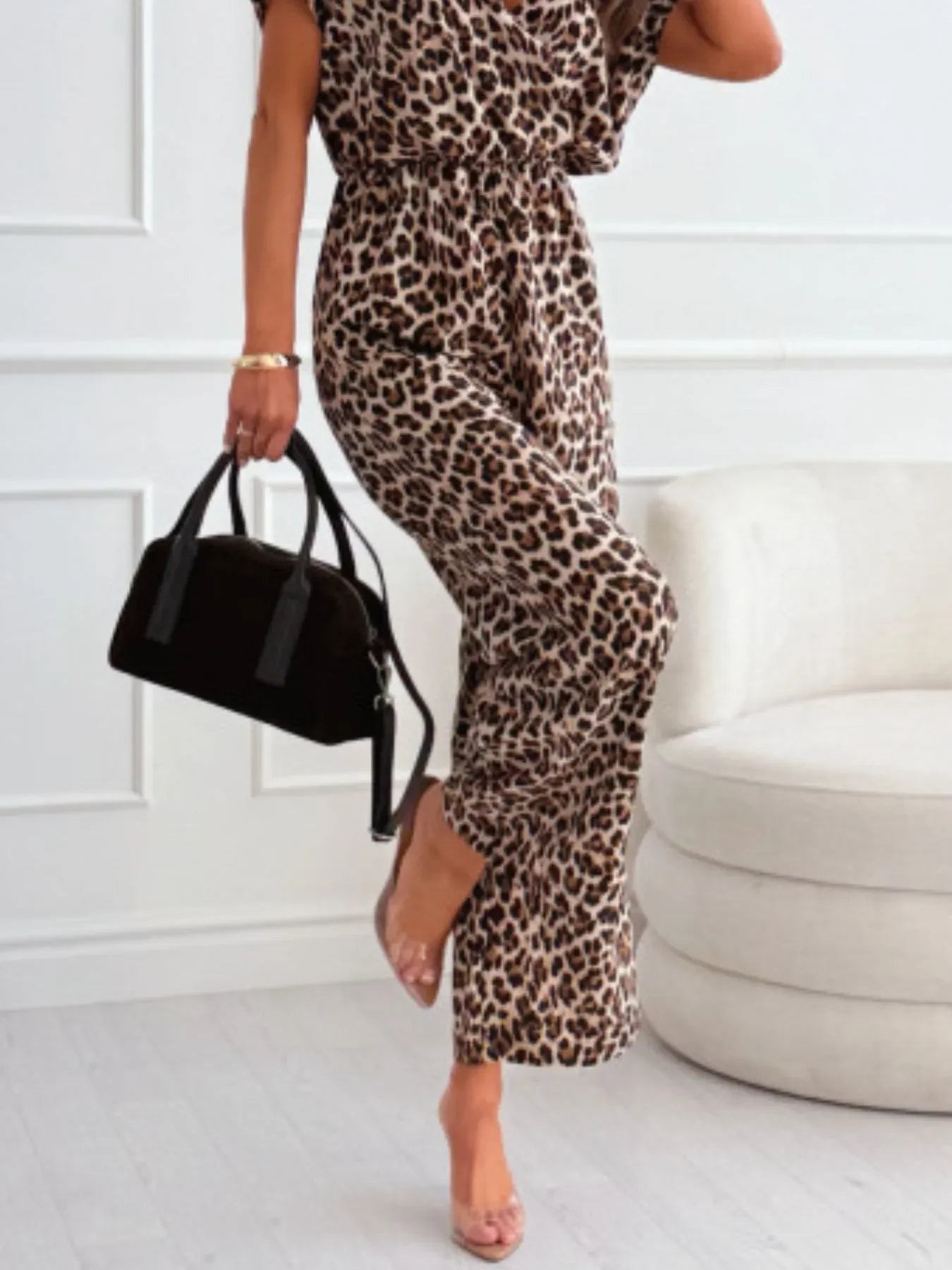 Woman wearing leopard print jumpsuit holding black handbag, standing indoors near white chair
