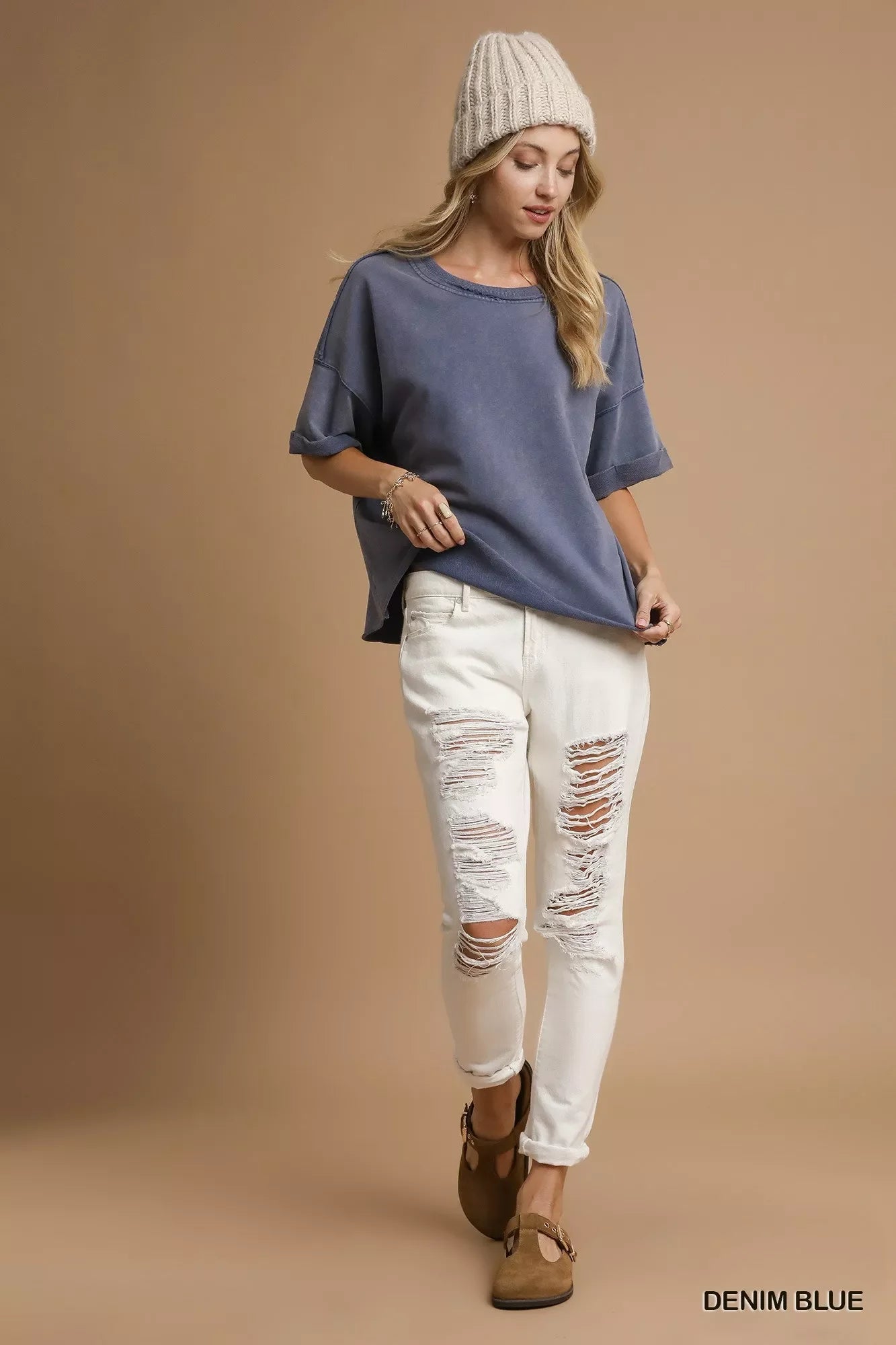 Woman wearing denim blue oversized top, ripped white jeans, knit beanie, and brown shoes on beige background