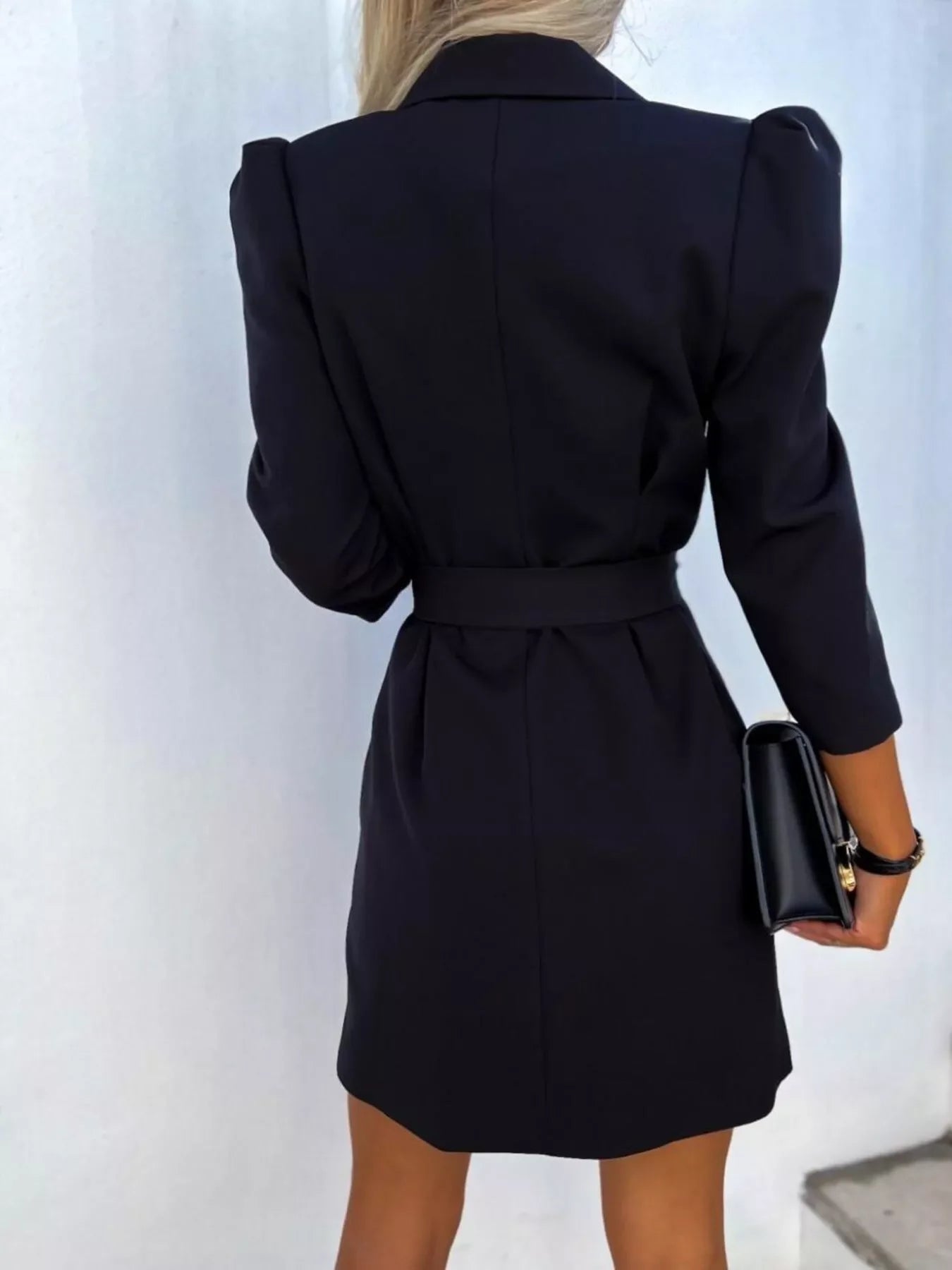Back view of woman in black belted blazer dress holding black clutch bag with white background