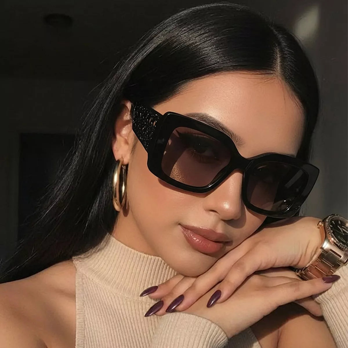 Woman wearing thick black square sunglasses, gold hoop earrings, and a beige ribbed top.