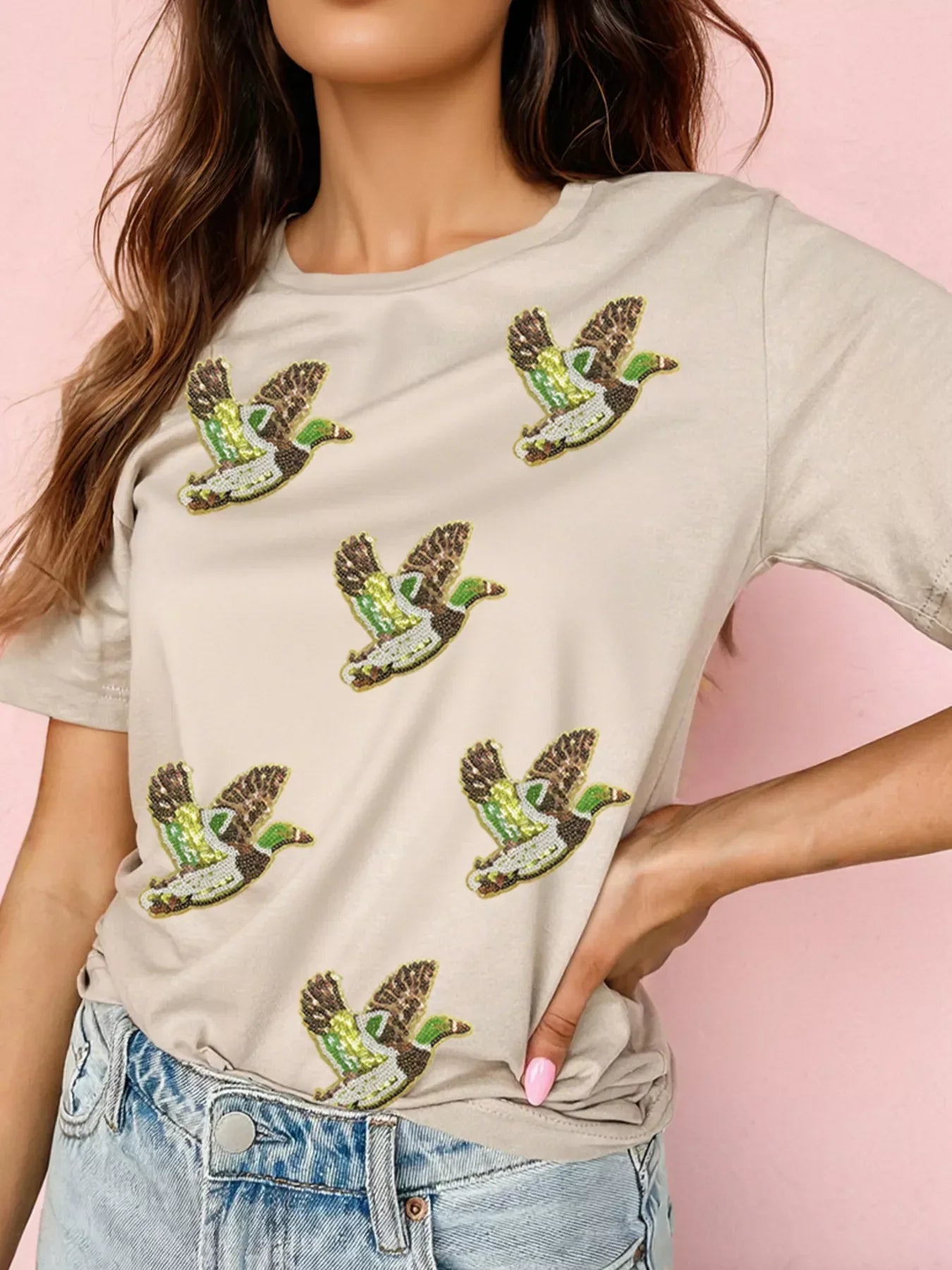 Beige t-shirt with sequined flying duck patches worn by woman with long hair, paired with light blue denim jeans