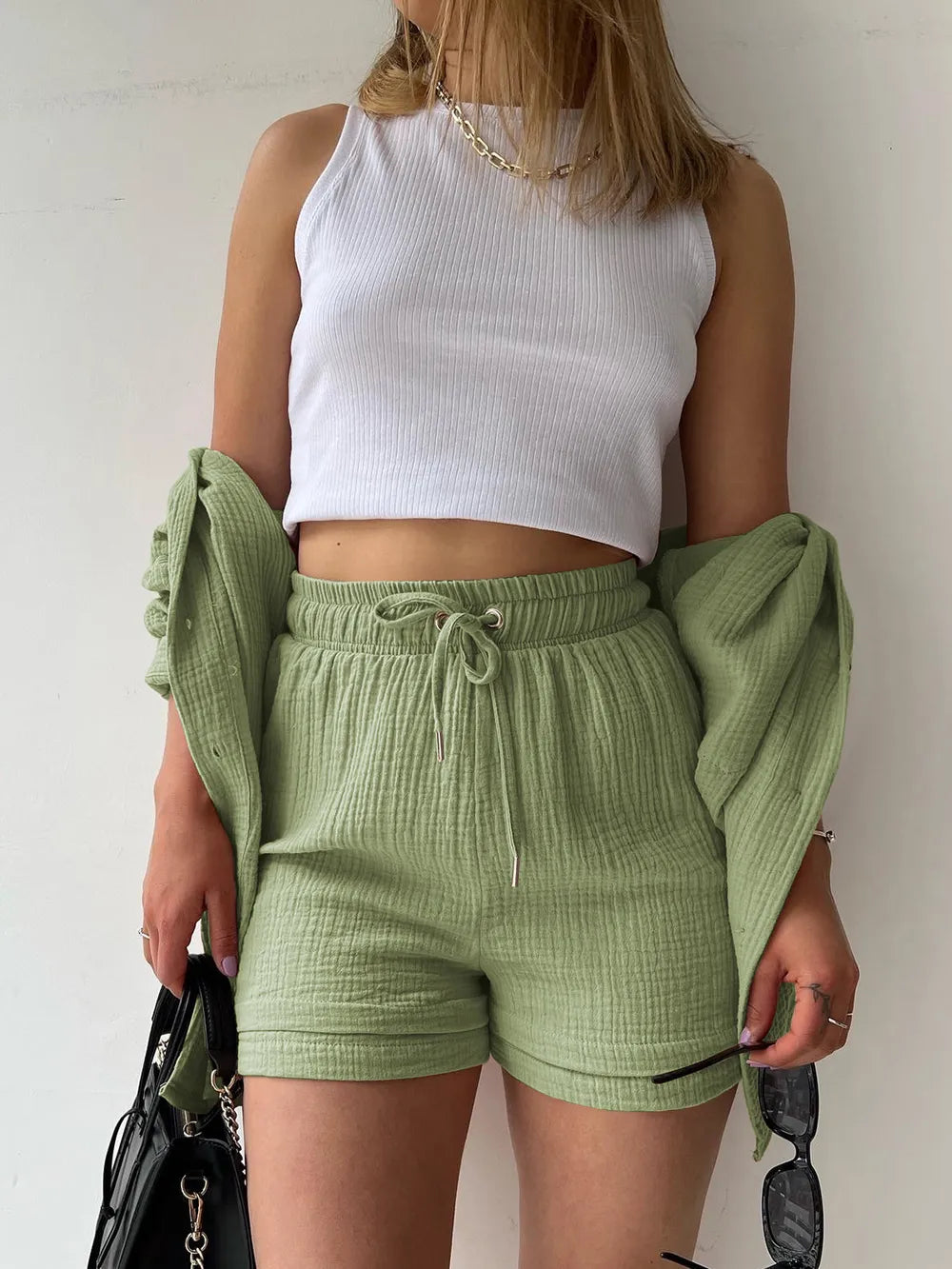 Woman in sage green shorts set and white ribbed crop top, holding a black handbag and sunglasses