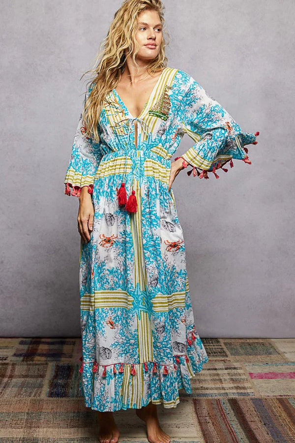 Woman wearing a boho maxi dress with blue and yellow coral print and red tassel accents