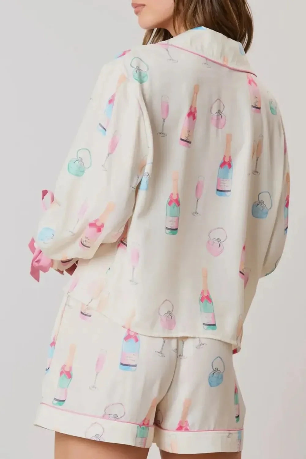 woman wearing cream pajamas with colorful champagne bottle, glass, and purse print, pink piping trim