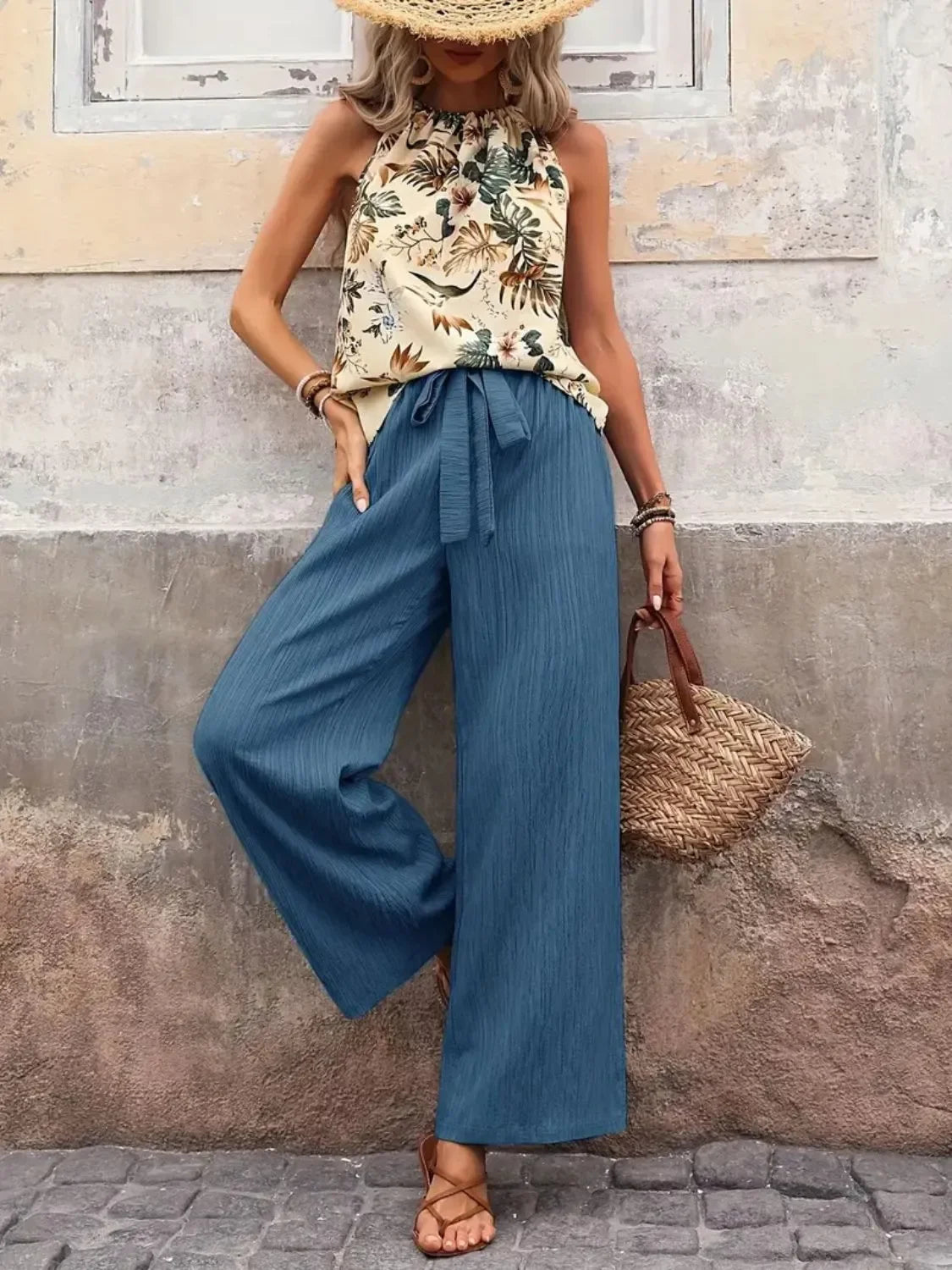 Woman wearing floral halter top, wide-leg blue pants, straw hat, and holding woven bag
