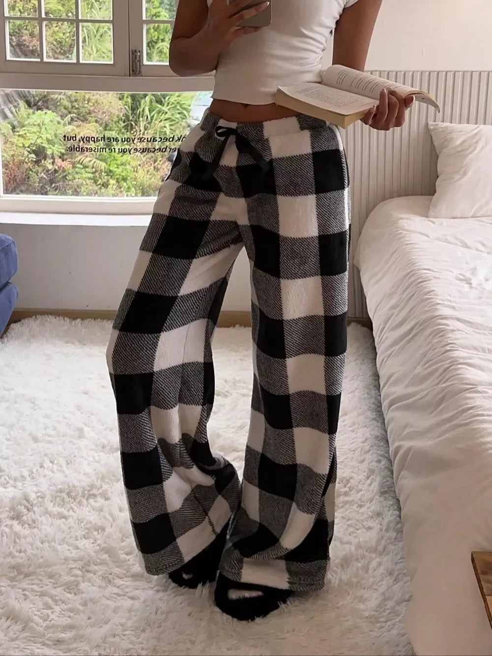 Woman wearing cozy black and white checkered wide-leg lounge pants and a white crop top, holding a book in a bright bedroom