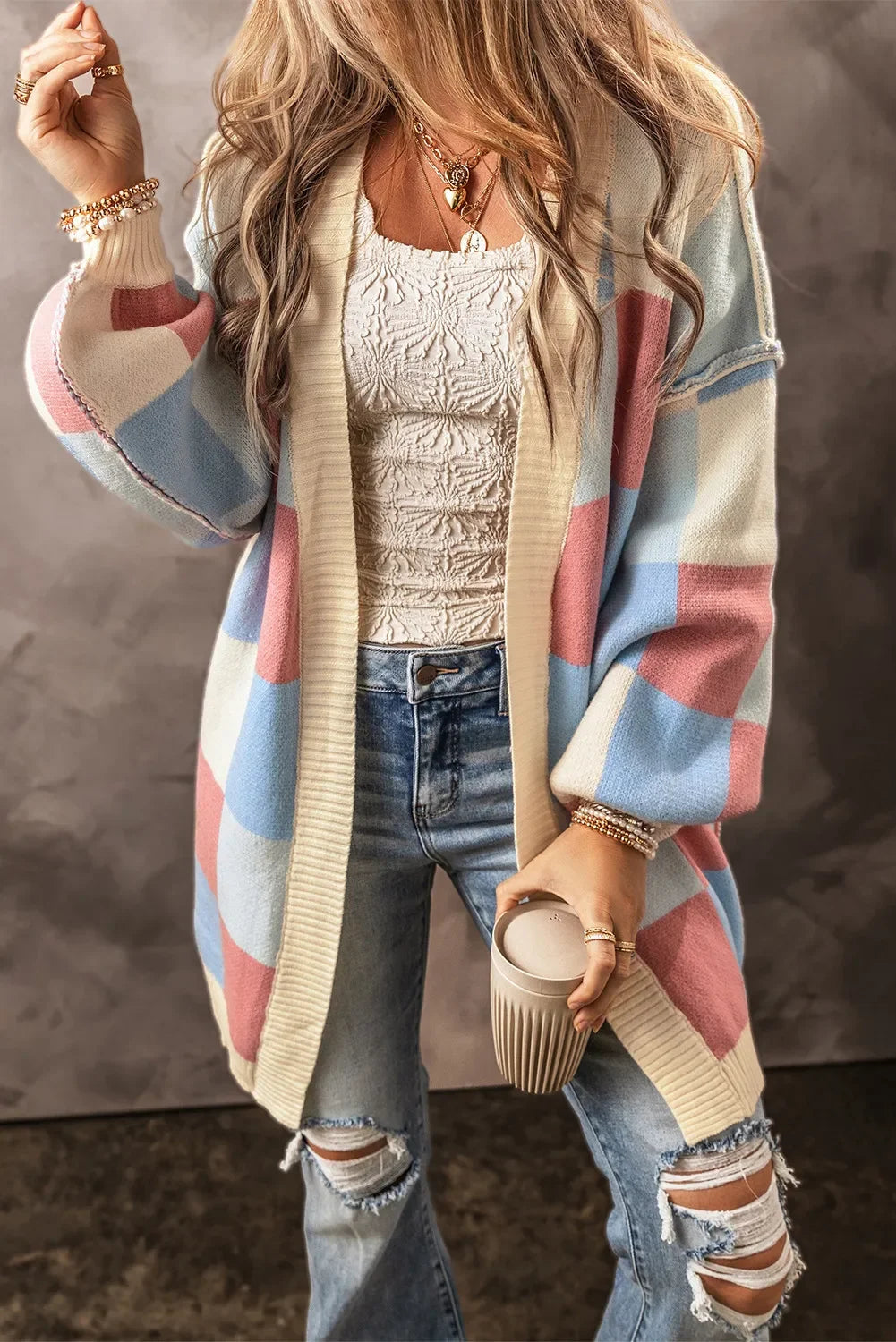 Woman wearing pastel checkered cardigan, white textured top, ripped jeans, holding beige coffee cup