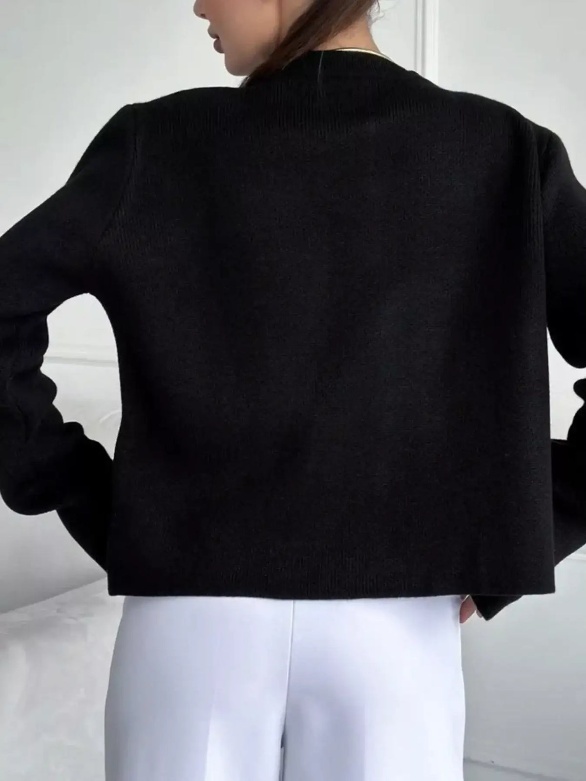 Back view of a woman wearing a black crew neck button down cardigan by Evie's Dreamland with white trousers.