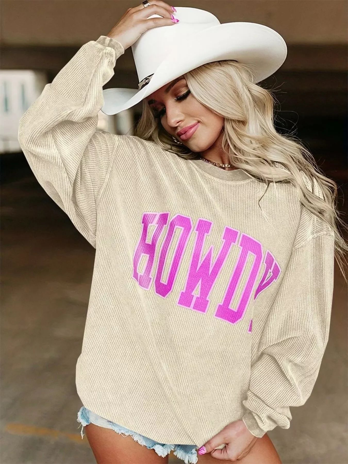 Blonde woman wearing beige corduroy sweatshirt with pink HOWDY text and white cowboy hat