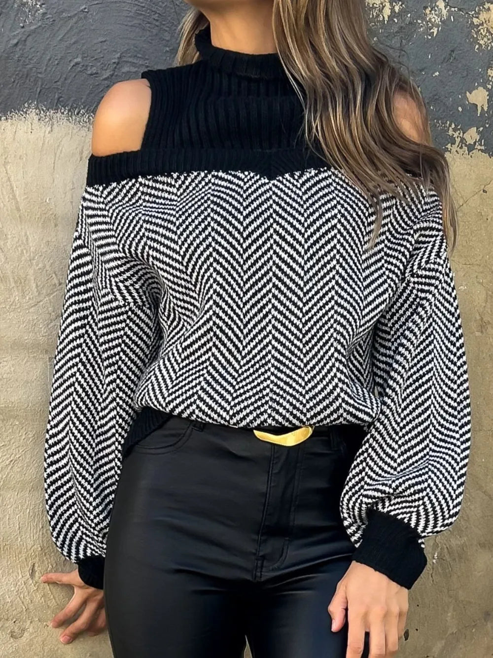 Woman wearing black and white chevron cold shoulder sweater with high neck and black pants against textured wall