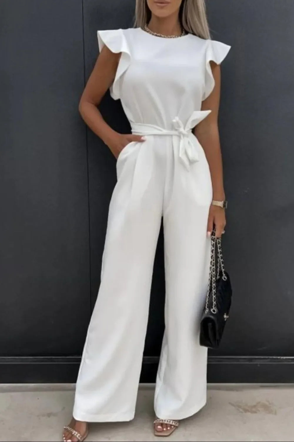 Woman wearing white ruffled sleeve jumpsuit with belt, holding black chain handbag, standing against black wall