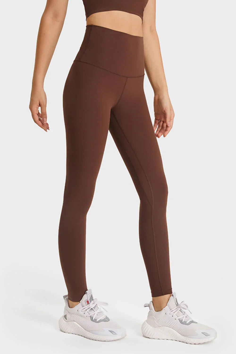 Woman wearing brown high-waisted leggings and white athletic sneakers on white background