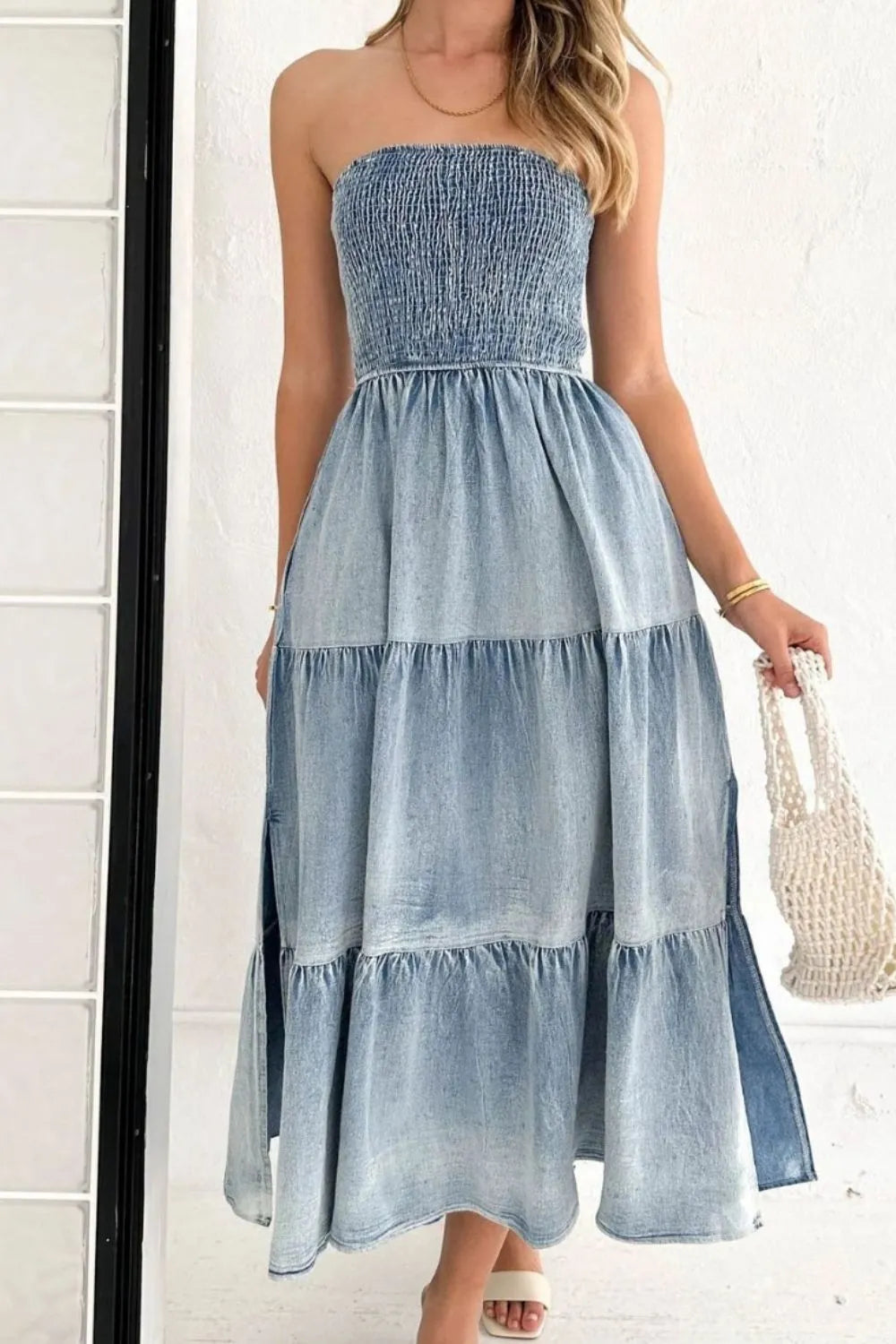 Woman wearing a strapless denim maxi dress with tiered skirt, holding a woven bag