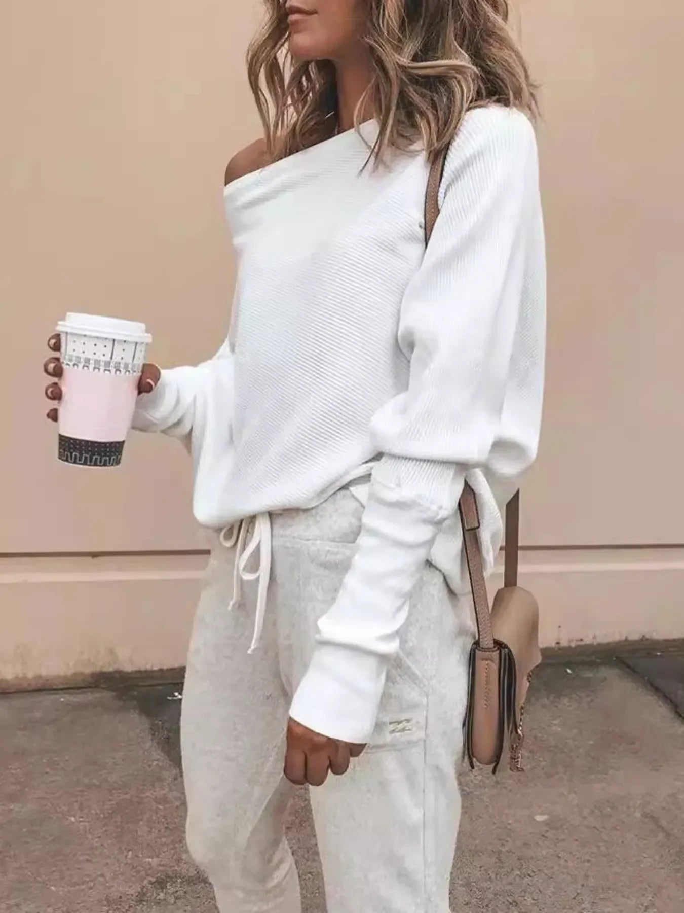 Woman wearing white off-shoulder sweater and light gray joggers holding coffee cup, beige crossbody bag
