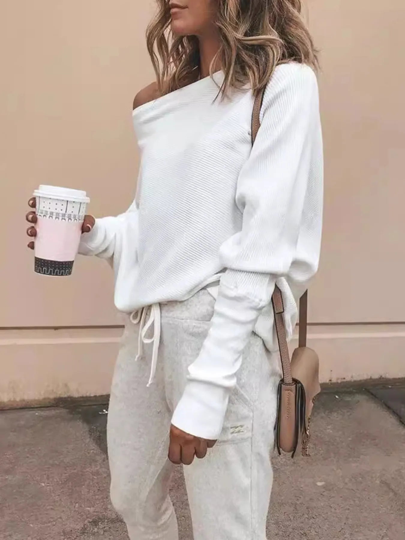 Woman wearing white off-shoulder sweater and light gray joggers holding coffee cup, beige crossbody bag