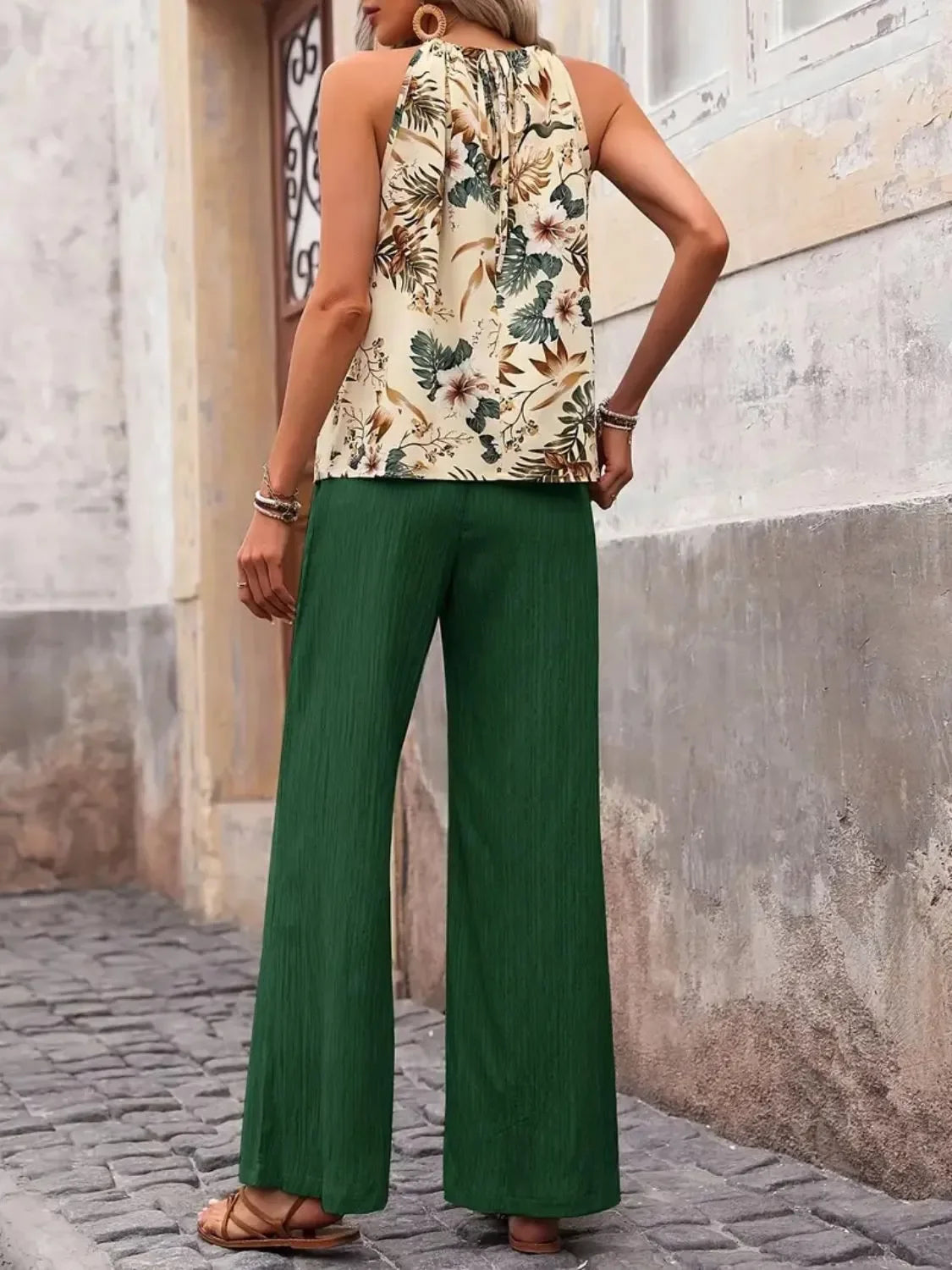 Woman wearing a sleeveless floral blouse and green wide-leg pants, standing on a cobblestone street.
