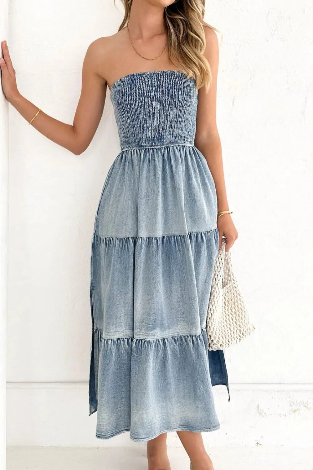 Woman wearing a strapless denim midi dress with a smocked bodice and tiered skirt, holding a white woven handbag