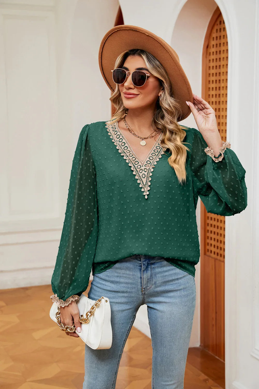 Woman wearing green lace V-neck blouse with balloon sleeves, light blue jeans, tan hat, sunglasses, and holding white handbag