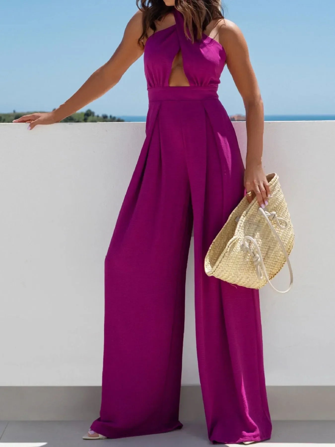 Woman in sleeveless purple wide-leg jumpsuit holding woven straw handbag outdoors