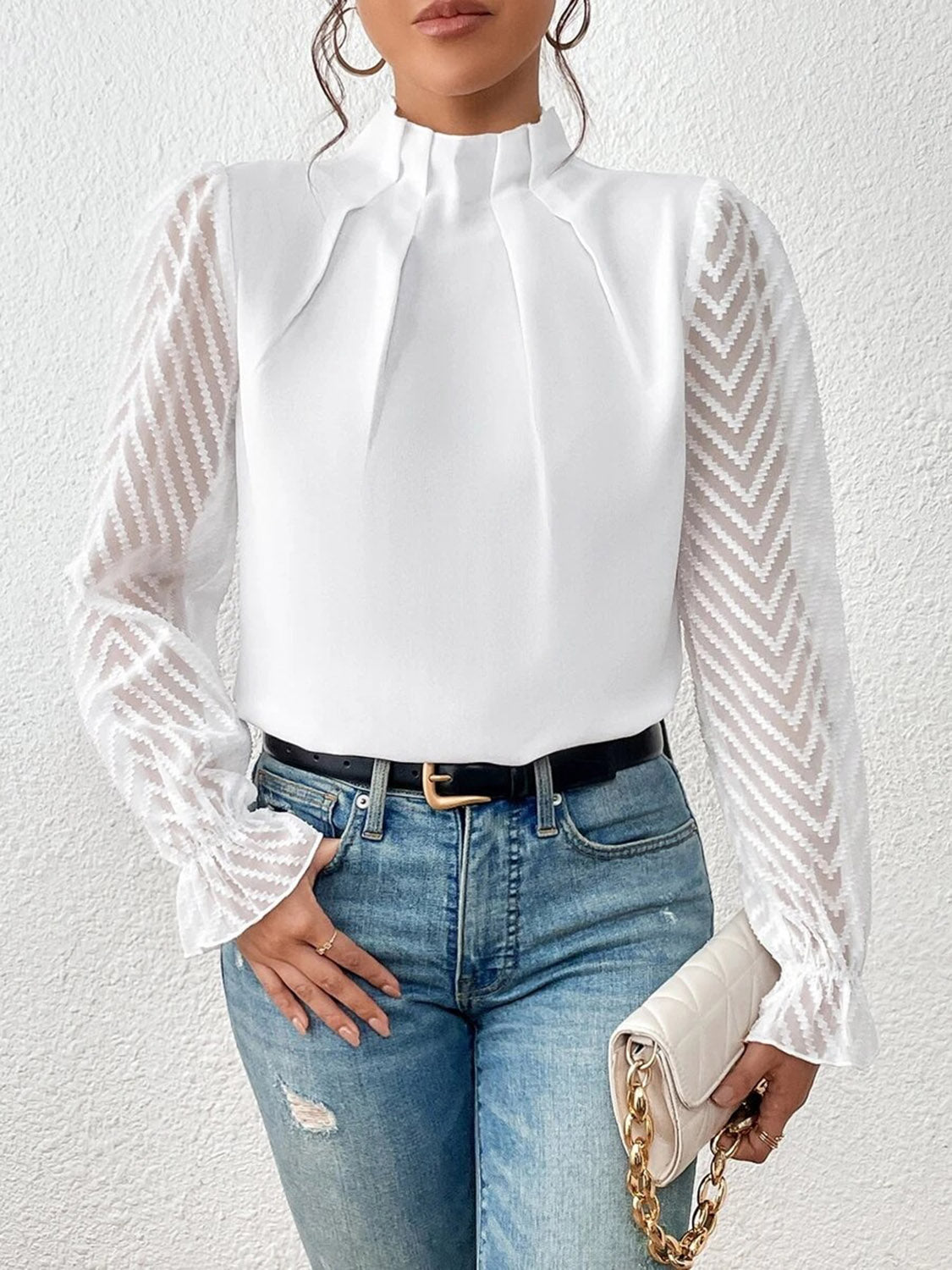 Woman wearing a white blouse with sheer chevron sleeves, blue jeans, and holding a quilted handbag.