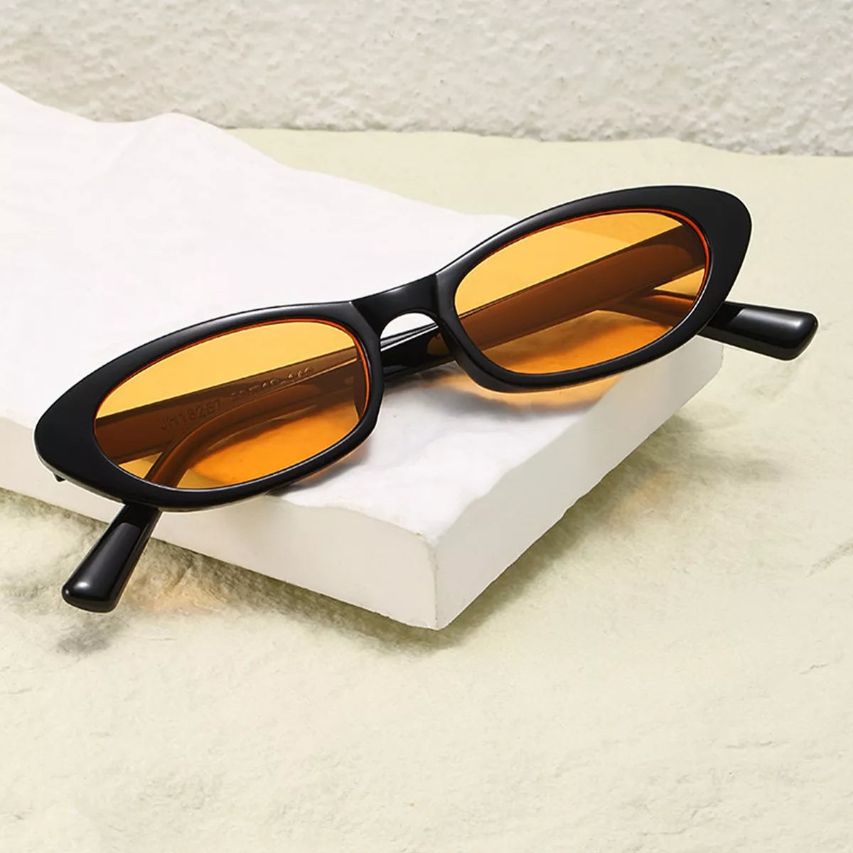 Black oval sunglasses with orange tinted lenses resting on a white textured surface.