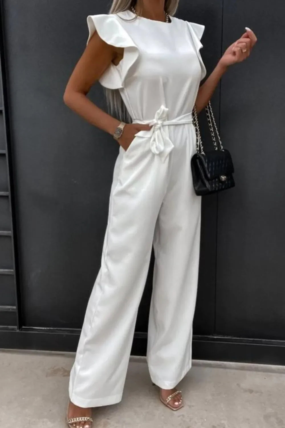 Woman wearing white ruffle sleeve jumpsuit with belt, holding black chain handbag, standing by black wall