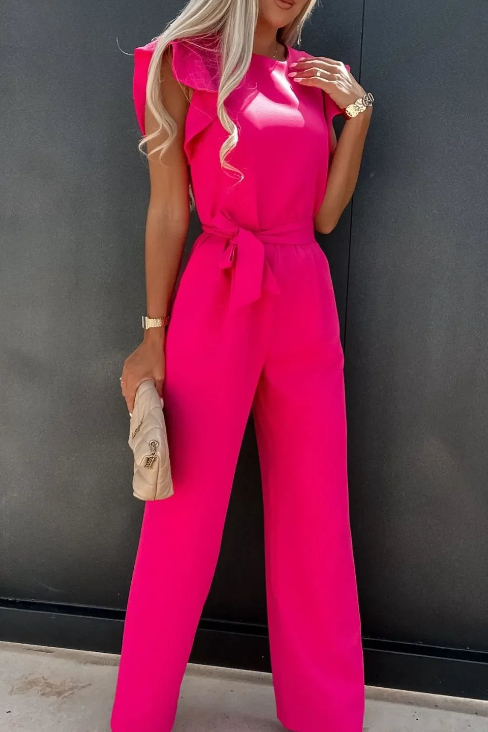 Woman wearing a bright pink sleeveless jumpsuit with a fabric belt, holding a beige quilted clutch