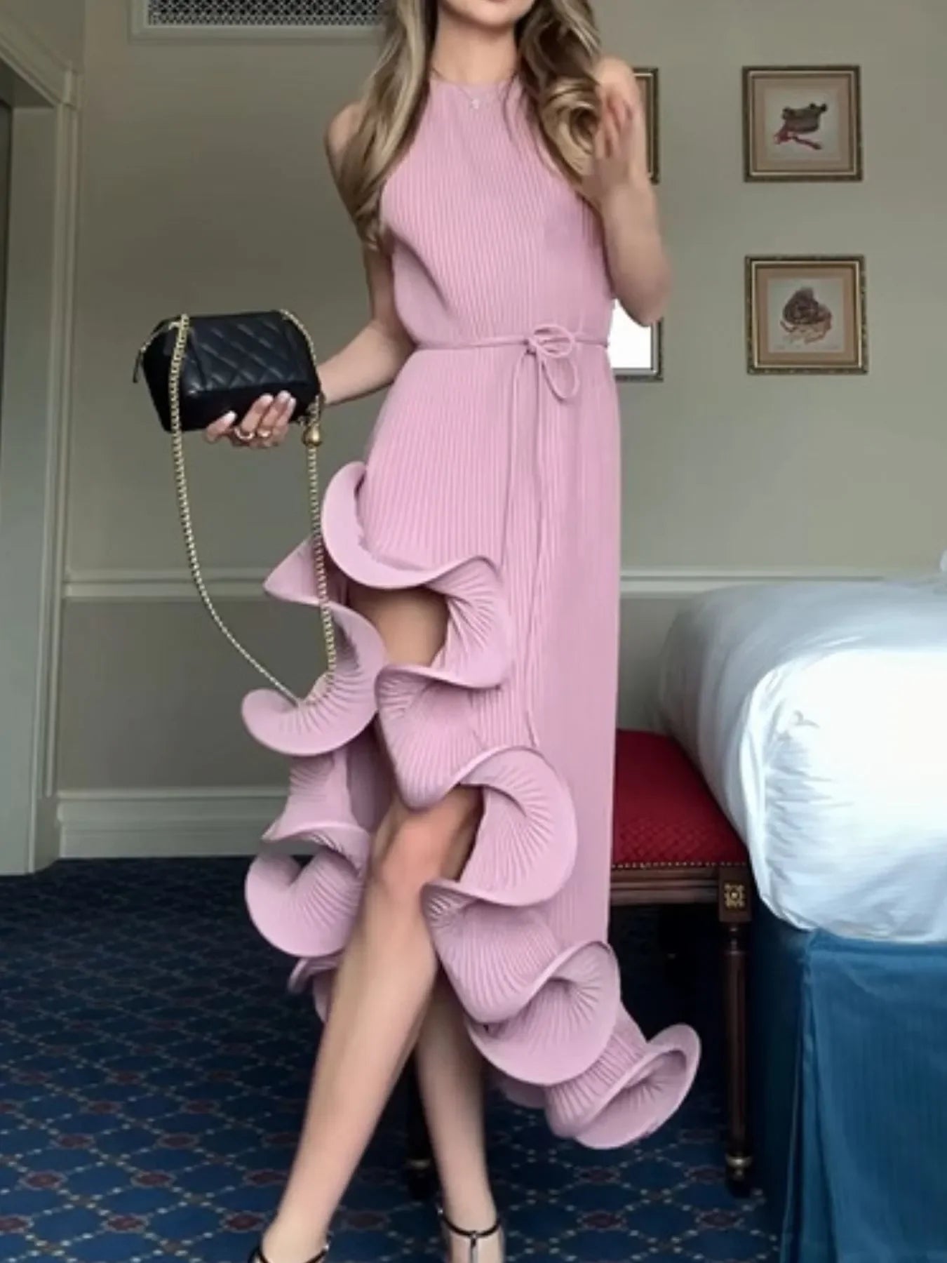 Woman in pink ruffle pleated dress holding black quilted purse in a stylish room