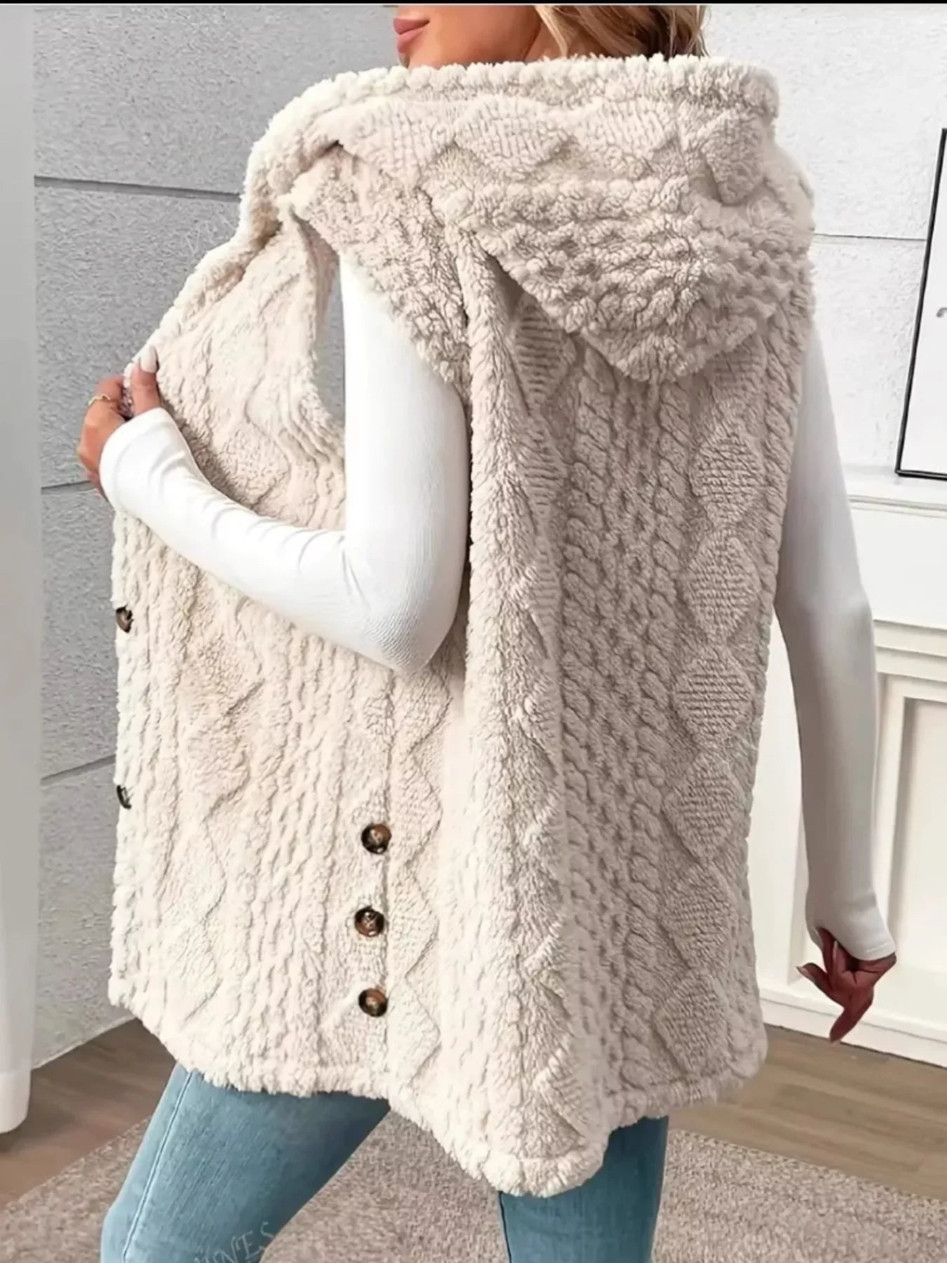 Woman wearing a beige knitted sleeveless hooded buttoned vest with textured patterns over white long sleeve top and blue jeans