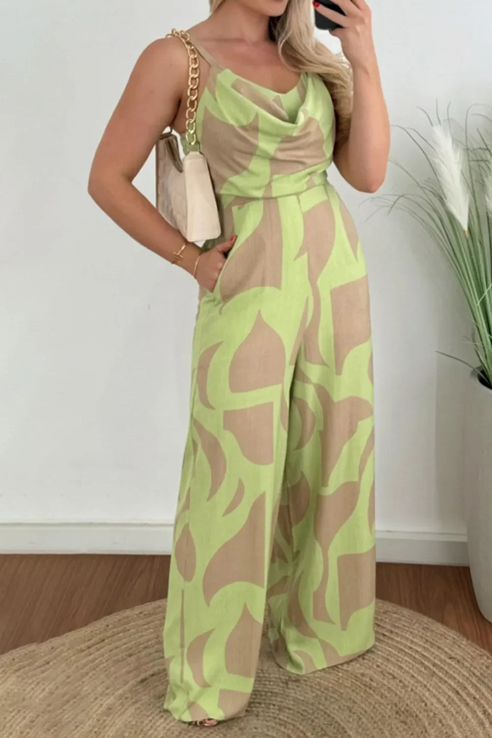 Woman wearing green and beige abstract print wide-leg jumpsuit with cowl neckline and holding beige bag