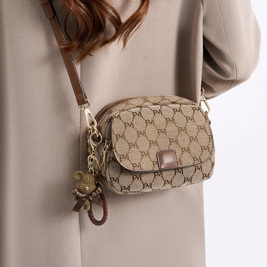 Woman wearing a beige patterned crossbody bag with brown leather strap and bear keychain.