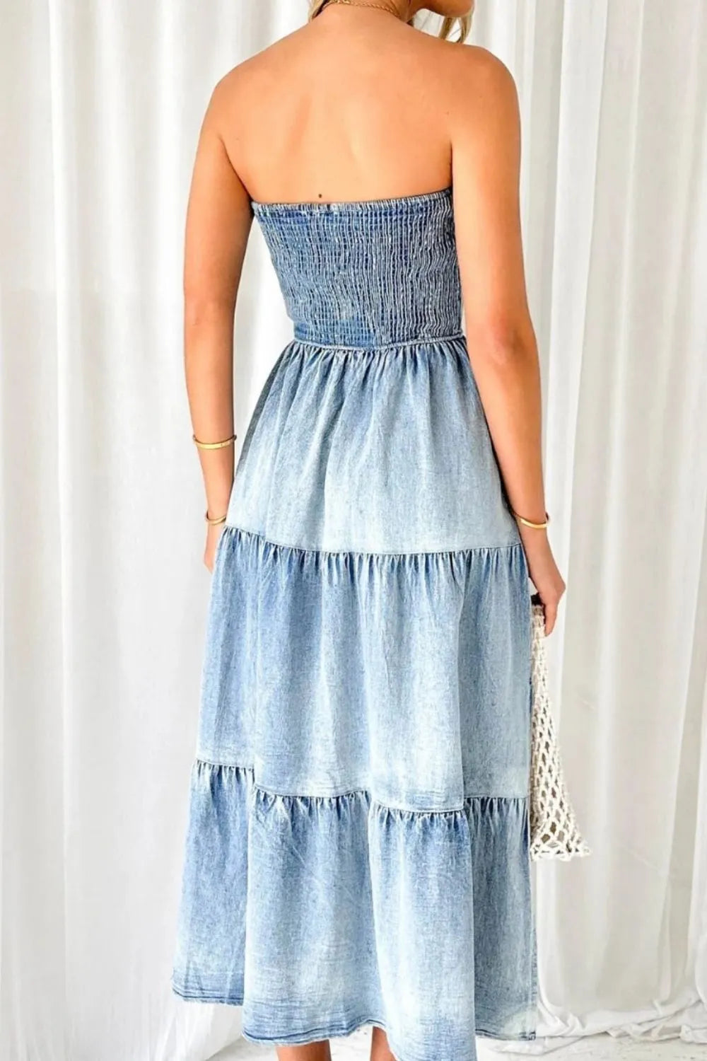 Woman wearing strapless tiered denim midi dress, holding woven bag, indoor setting