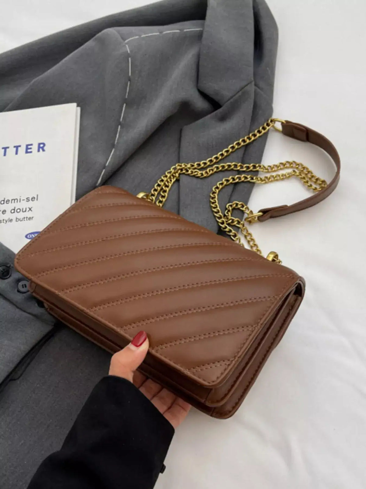 Brown quilted flap chain shoulder bag by Evie's Dreamland, held against a grey fabric backdrop.