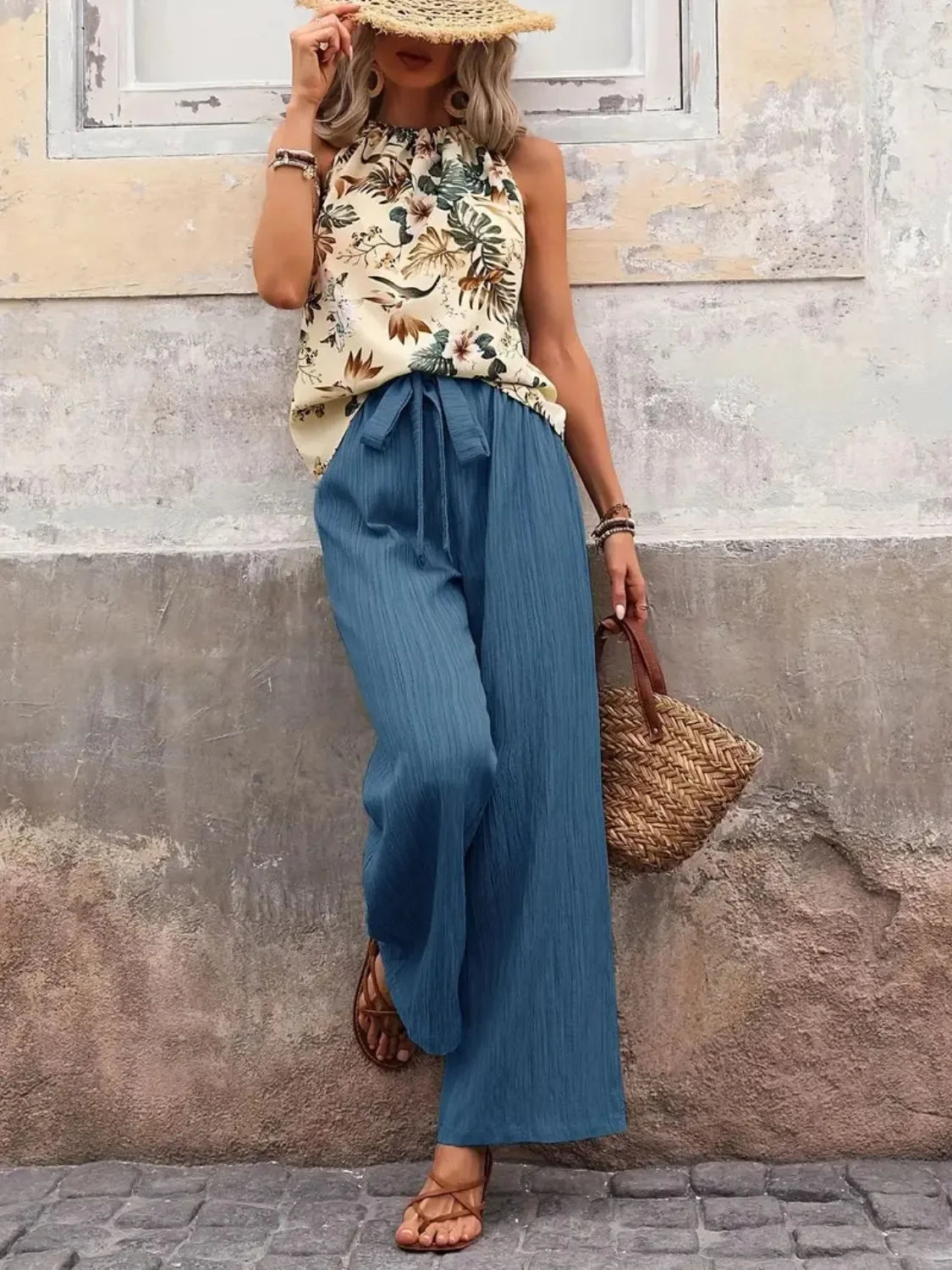 Woman wearing a floral sleeveless top with blue wide-leg pants and straw hat, holding a woven bag