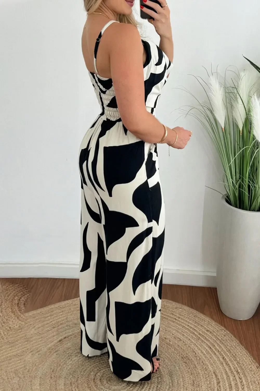 Woman in black and white abstract print wide-leg jumpsuit posing indoors near tall green plant