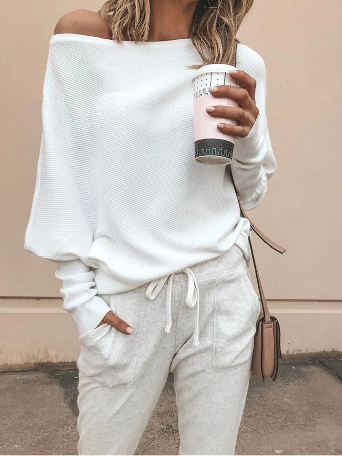 Woman wearing off-shoulder white sweater and light gray sweatpants holding a patterned coffee cup