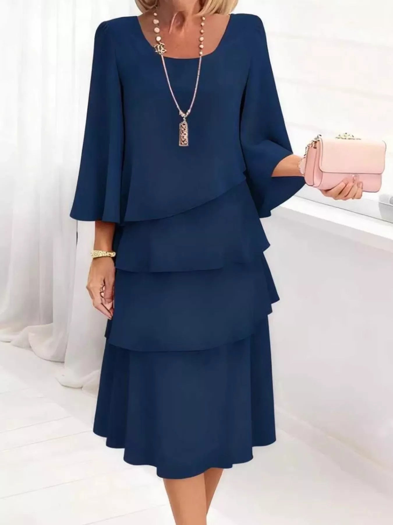 Navy blue tiered midi dress with bell sleeves, styled with pink clutch and pearl necklace for elegant occasions.