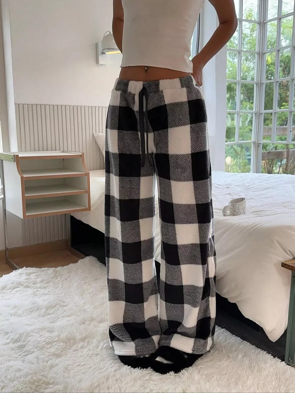 Woman wearing cozy black and white checkered fleece lounge pants and white cropped tank top in a bright bedroom